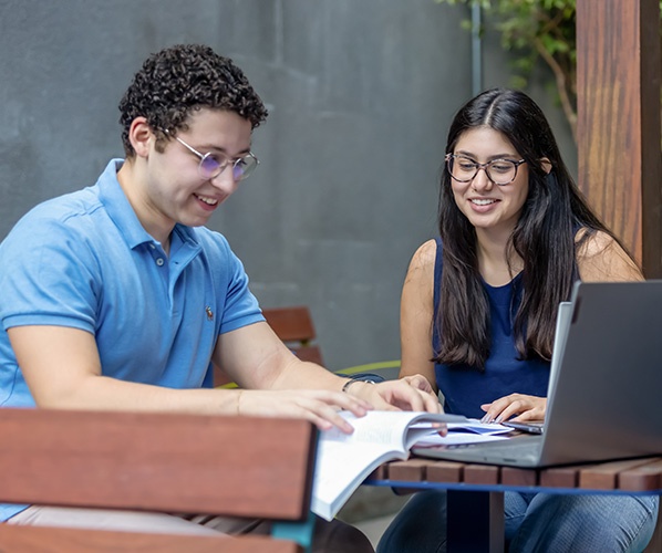 casal de alunos estudando juntos numa mesa com computador e livros.